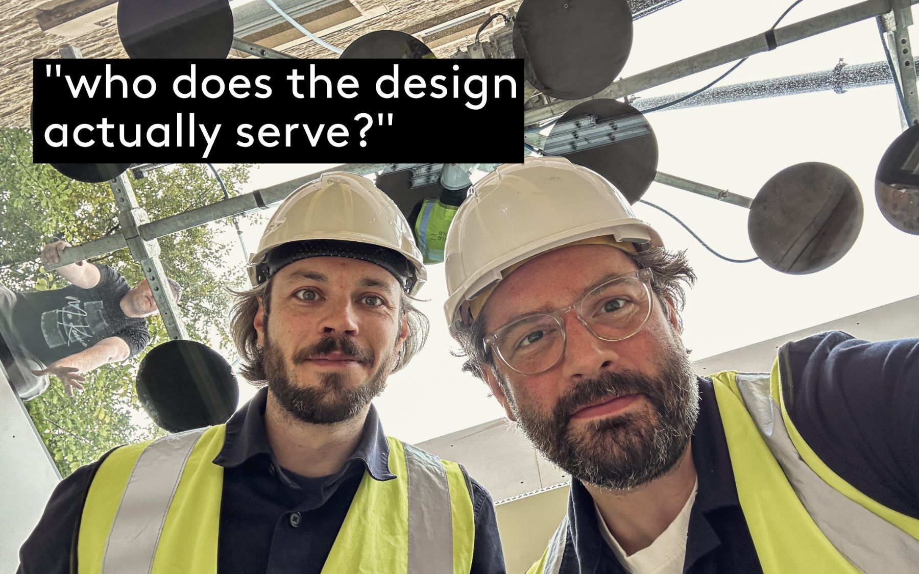 Andrew and Jack in hard hats and hi-vis on a scaffolded site, looking up at the camera beneath the scaffold deck