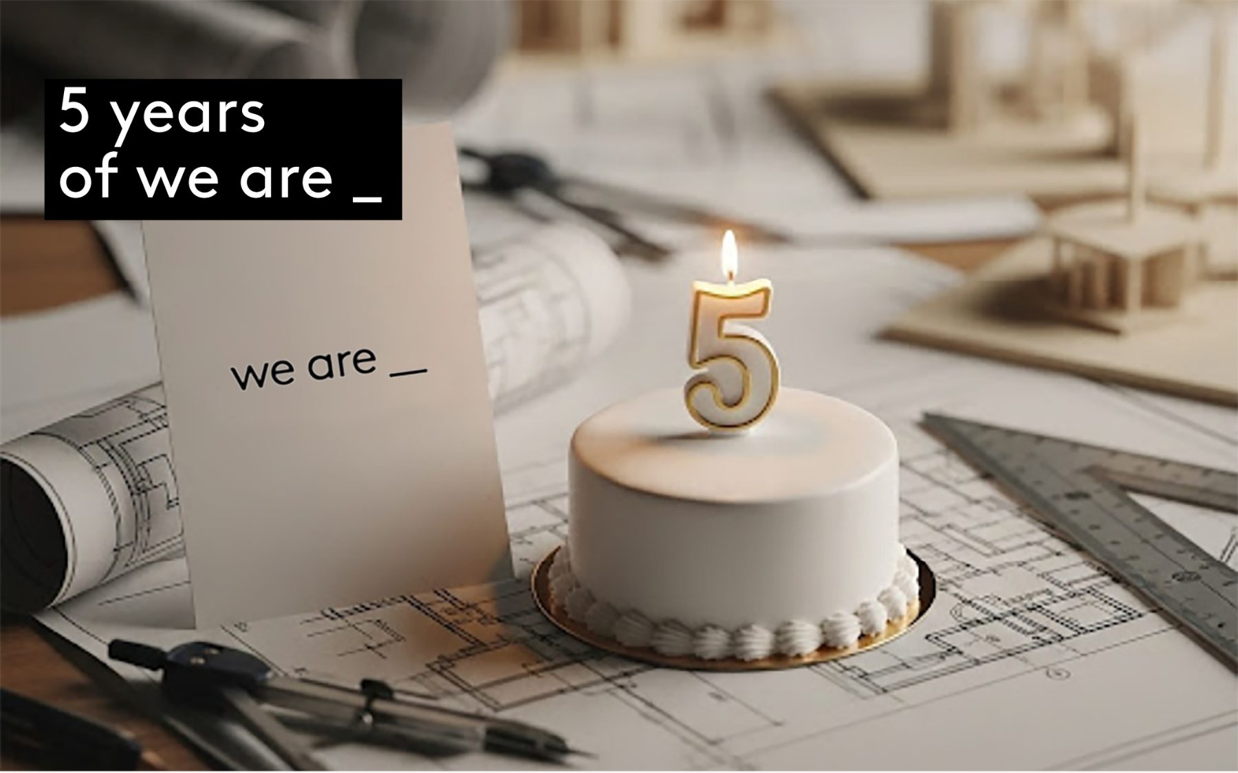 A white celebration cake with a gold number five candle, beside a we are _ card on architectural drawings in the studio